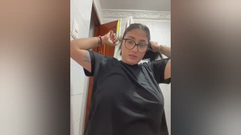 Video of a middle-aged woman with medium skin tone, wearing glasses and a black T-shirt, standing in a bathroom with white tiles and a wooden door.
