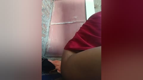 Video of a woman's thigh in a red skirt, partially obscured by a pink brick wall. Background shows a cluttered room with a brick wall and a bed with a pink blanket.