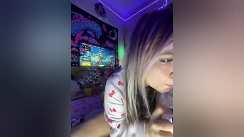Video of a young woman with long, straight blonde hair in a white shirt with red hearts, playing a video game in a colorful, lit room with a large TV displaying an anime scene.