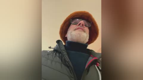 Video of an older man with a white beard, wearing a brown hat, dark glasses, and a grey quilted jacket with a black turtleneck underneath. He stands against a blurred beige wall.