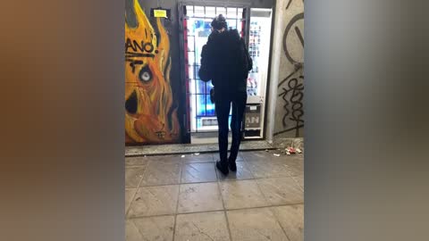 Video of a person in a black hoodie and pants standing at a door with graffiti of a fiery face. The floor is tiled, and the background shows a cluttered, graffitied wall.