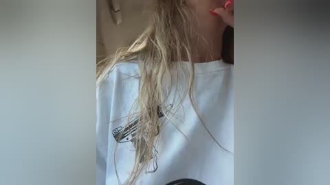 Video of a young woman with long, wet, blonde hair, wearing a white T-shirt with a black graphic design, and holding a black smartphone to her face, against a neutral background.