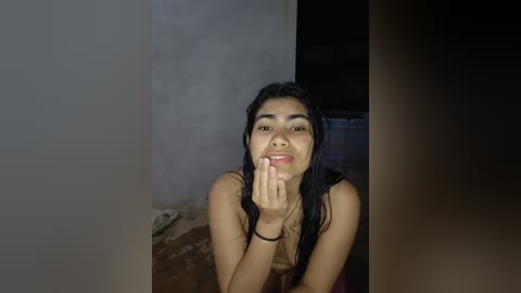 A young woman with long, wet black hair, wearing a black top, stands indoors against a dimly lit, textured wall. She has a medium complexion, and her expression is neutral.