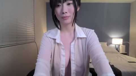 Media: A video of a fair-skinned Asian woman with black hair, wearing a semi-transparent white blouse, posing in a modern bedroom with beige walls, a bed, and a lamp.