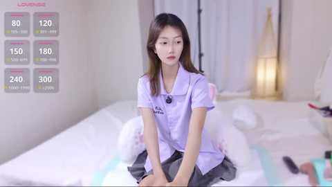 Media: Video of a young Asian woman in a light purple blouse and gray skirt sitting on a white bed in a softly lit, minimalist bedroom.