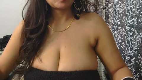 Media: Video of a topless woman with large breasts, wearing a black bandeau and gold chain necklace, in a room with floral-patterned curtains.