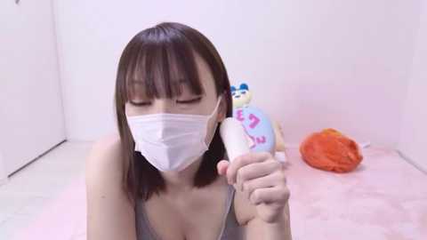 Media: Video of an East Asian woman with straight brown hair and light skin wearing a white surgical mask, holding a pink and blue plush toy, standing on a pink rug in a sterile, white-walled room.
