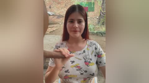 Media: A video of a young woman with long red hair, wearing a white Mickey Mouse t-shirt, sitting on a dirt floor, with a man's hand on her shoulder, in a rustic, outdoor setting.