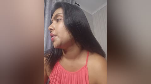 Media: Video of a young South Asian woman with long black hair, wearing a red sleeveless blouse, looking to the left, against a blurred background.