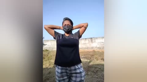 Video of a woman with medium skin tone, wearing a black face mask, grey t-shirt, and plaid shorts, stretching arms, outdoors near a concrete wall under a clear blue sky.