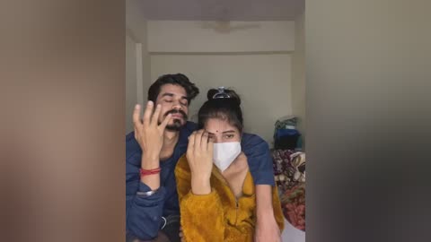 Video of a man with a beard and a woman with a bun, wearing masks, standing in a dimly lit room with white walls and a ceiling fan.