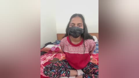 A video of a South Asian woman with long black hair, wearing a red sweater, black face mask, and colorful pajamas, sitting cross-legged on a bed with colorful bedding in a simple room.