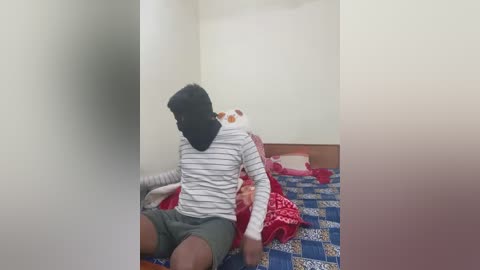 Video of a person in a striped shirt and green shorts sitting on a blue and white checkered bed, with a white stuffed toy and red blanket behind them.