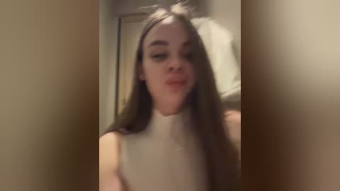 Video of a young woman with long brown hair, wearing a white sleeveless top, making a pouty face. The background is blurred, featuring beige walls and a partially visible white pillow.