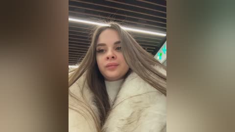 Video of a young woman with long, straight brown hair, wearing a white turtleneck sweater and a beige fur coat, indoors with a dark wooden ceiling and bright fluorescent lights in the background.
