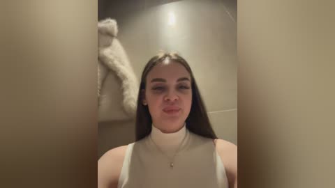 A video of a young woman with long dark hair, wearing a white sleeveless top, standing in a narrow hallway with beige walls. A stuffed animal is hanging on the left wall.