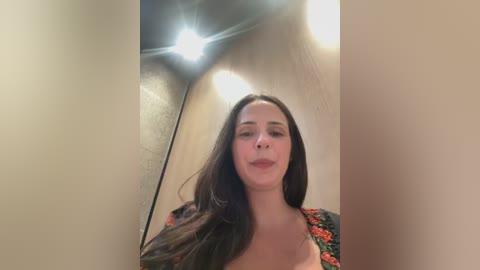 Video of a young woman with long dark hair, light skin, and a neutral expression, wearing a black floral top. The image is taken in a dimly lit room with a bright overhead light source.