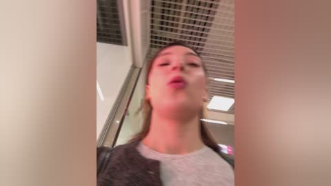 Media: A blurry, low-angle video of a young woman with light skin and brown hair, wearing a gray sweater and black jacket, indoors with a metallic ceiling and fluorescent lights.