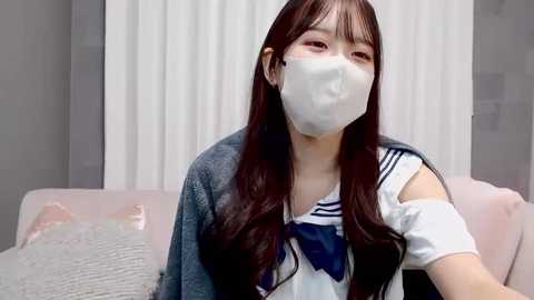 A video of a young Asian woman with long, straight black hair, wearing a white mask, a blue sailor collar top, and a gray cardigan, seated indoors.
