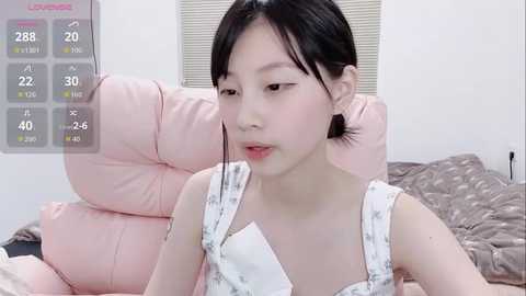 Video of a young East Asian woman with short black hair, wearing a white floral-patterned top, sitting on a pink couch in a minimalist bedroom.