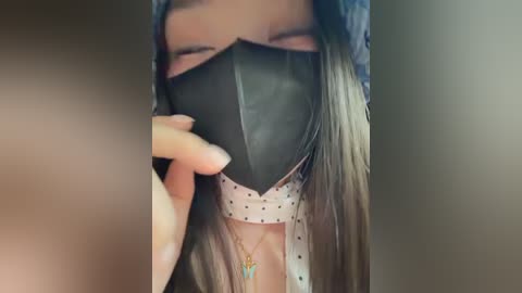 A close-up video of a person wearing a black paper mask with polka dots, covering their nose and mouth. The background is blurred, highlighting the mask's texture and the person's light brown hair.