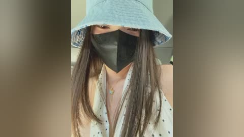 A video of an Asian woman with long, straight brown hair, wearing a light blue bucket hat, black face mask, and a white shirt with polka dots. The background is blurred, featuring neutral tones.