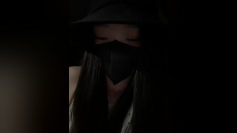 A dimly lit video of a person with a black hood, face mask, and dark clothes, giving a mysterious, shadowy appearance against a dark background.
