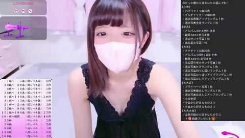 Video of an East Asian woman with straight, shoulder-length brown hair, wearing a black lace-trimmed top and a white face mask, sitting in a room with pastel-colored walls and a desk. Text overlay shows Japanese chat messages.