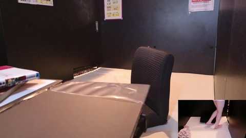 Video of a dimly lit, minimalist office with dark walls, a black cushioned chair, a closed laptop, and scattered papers. A close-up inset shows a person's hand holding a pen over a document.