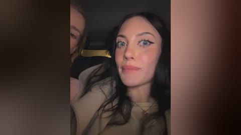 A video of a young woman with pale skin, blue eyes, and long dark hair, wearing a beige sweater, standing next to a person with a brown mask, in a dimly lit room.