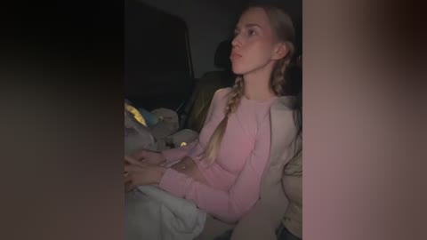 A dimly-lit video of a young, pale-skinned woman with blonde braids, wearing a light pink sweater, sitting in a car at night.