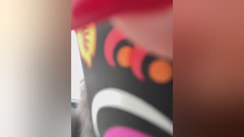 A blurry, close-up image of a person with pink hair and a black and white outfit, featuring orange and yellow accents. The background is indistinct, suggesting a possible indoor setting.