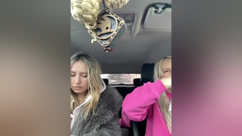 Video of two blonde women in a car, one in a fur coat and the other in a pink jacket, with a large, intricate decorative piece hanging from the ceiling.