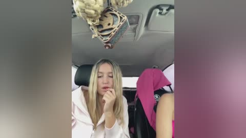 Video of a blonde woman in a white robe, hand on her chin, seated in a car with a decorative skull hanging from the ceiling, surrounded by pink and black luggage.