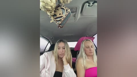 Video of two young women with long blonde hair, one in a pink jacket, the other in a pink strapless dress, seated in a car, looking tired and bored.
