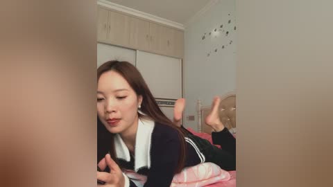 Video of an Asian woman with long brown hair, wearing a black and white school uniform, lying on a pink bed, smiling, with a blurred figure in the foreground.