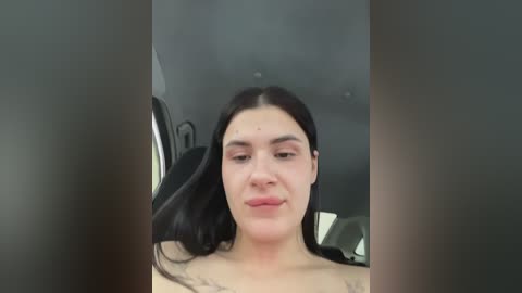 Media: A video of a young woman with long black hair, fair skin, and small breasts, sitting in a car. Her expression is neutral, and she's wearing a black top. The car's interior is visible in the background.