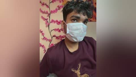 Video of a young South Asian man with short, curly hair and a light brown complexion, wearing a maroon shirt with a deer graphic and a blue surgical mask. Background features pink cherry blossom wallpaper.