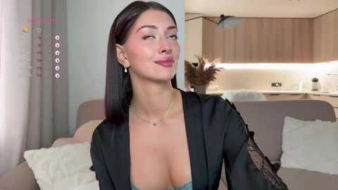 Video of a young woman with light skin and straight dark hair, wearing a black lace robe, smiling in a modern living room with a gray couch and wooden cabinets.