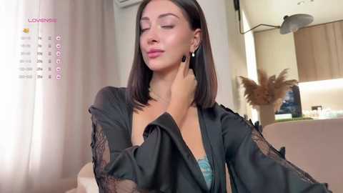 Video of a young woman with straight black hair, wearing a black lace robe, touching her neck, in a modern, softly lit living room.