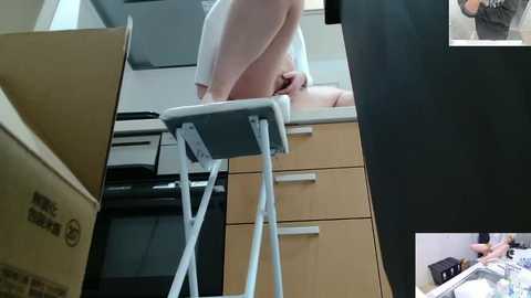 Video of a nude person standing on a chair in a modern kitchen, with a cardboard box in the foreground and a person in the background holding a camera.