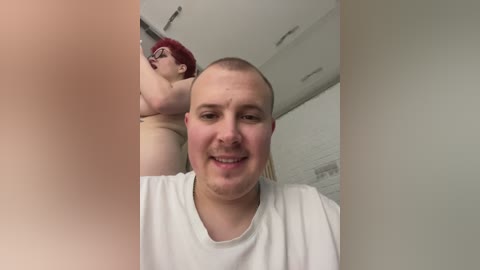 A video features a bald, grinning man in a white T-shirt. Behind him, a woman with red hair, glasses, and a tattooed arm is seen in a bathroom.