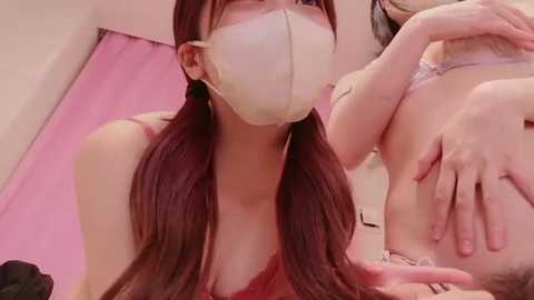 Video of a young woman with long red hair, wearing a face mask, leaning over a partially visible man on a pink bed, holding his hand.