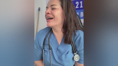 Video of a smiling, middle-aged woman with medium-length brown hair, wearing a blue scrub top and stethoscope, standing in a clinical setting with a blue poster and medical equipment in the background.