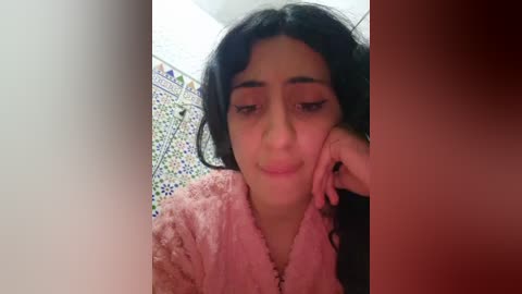A video of a young woman with long black hair, light skin, and brown eyes, wearing a pink fuzzy robe, leaning on a tiled bathroom wall with intricate patterns.