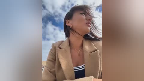 Video of a young Asian woman with long black hair, wearing a beige coat, looking upward against a blue sky with scattered clouds.