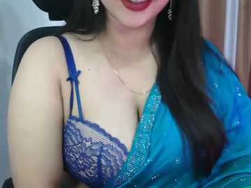 Video of a South Asian woman with fair skin, long dark hair, and red lipstick, wearing a blue lace bra and a turquoise saree, seated in a black chair.