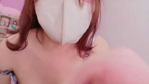 Video of a light-skinned woman with long brown hair, wearing a white heart-shaped mask, revealing a bare shoulder and upper chest. Background features a pink room with toys and shelves.