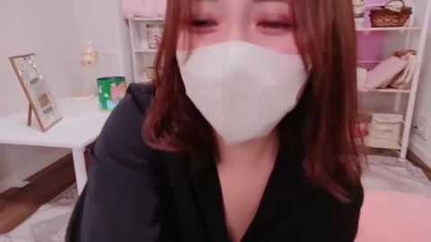 Video of a woman with medium-length, straight brown hair, wearing a white mask, black top, and sitting in a room with a white table, green cup, and pink shelves.