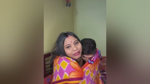 Video of a smiling woman with dark hair, wearing a colorful orange and pink blanket, holding a child with dark hair, in a simple, brightly lit room with yellow walls.